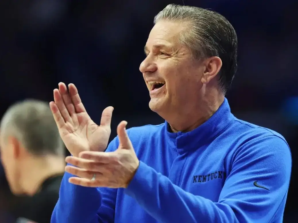 John Calipari coaching courtside in a suit with cheering fans in the background during a college basketball game